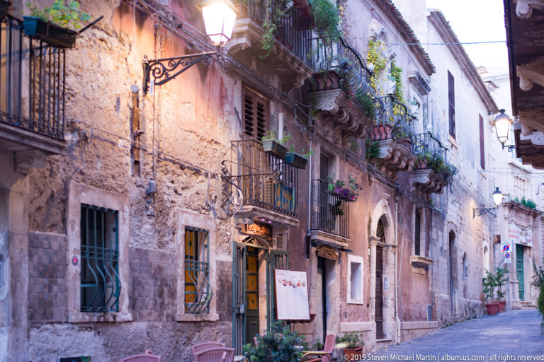 Narrow Ortigia lane with weathered stone façades, wrought-iron balconies, potted plants, shutters, and outdoor seating.