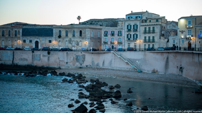 Historic buildings and glowing street lamps above a high seawall enclosing a small rocky cove on Ortigia.