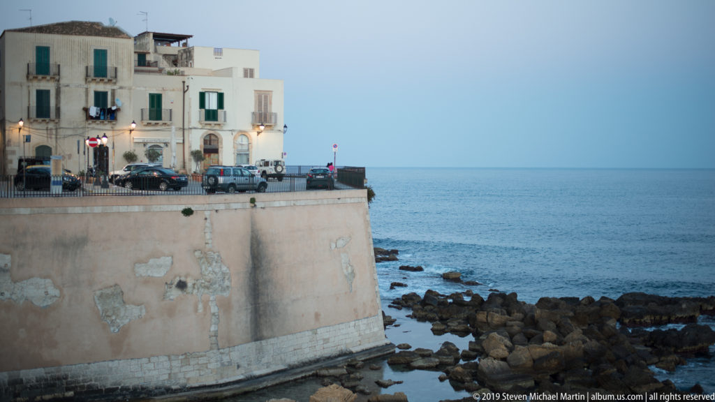 Tall pale masonry seawall above a rocky shore, topped by historic Ortigia buildings, street lamps, and parked vehicles.