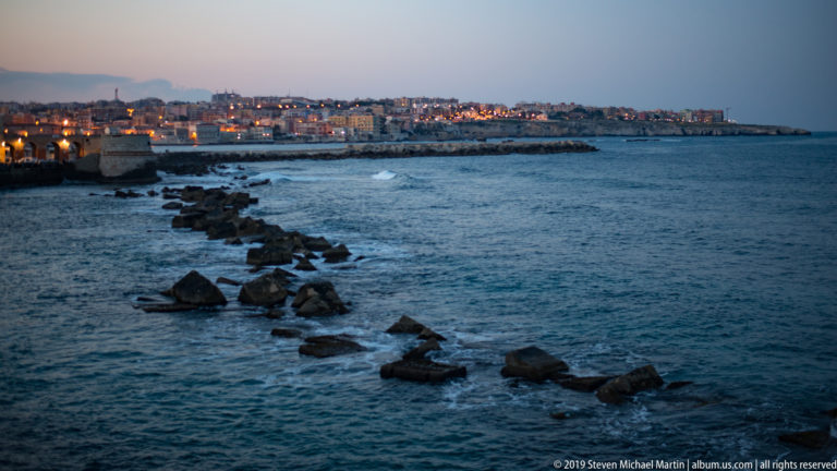 Large dark rocks form a diagonal nearshore barrier in blue water, with illuminated Ortigia buildings in the distance.