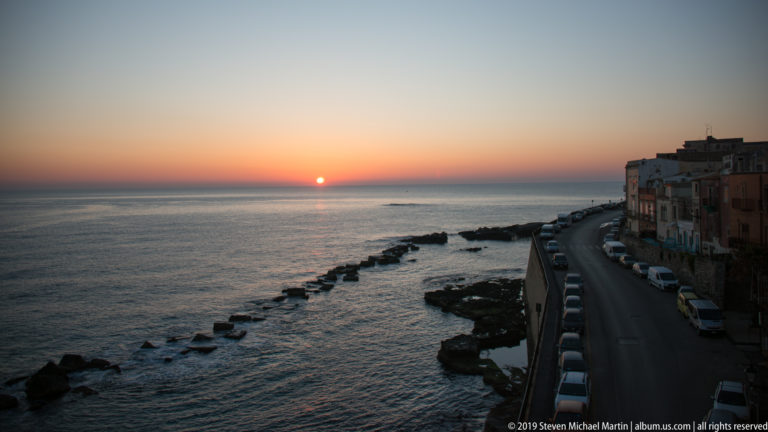 Peach-colored sunset over the sea with rocks and a curving Ortigia waterfront road lined by parked cars.