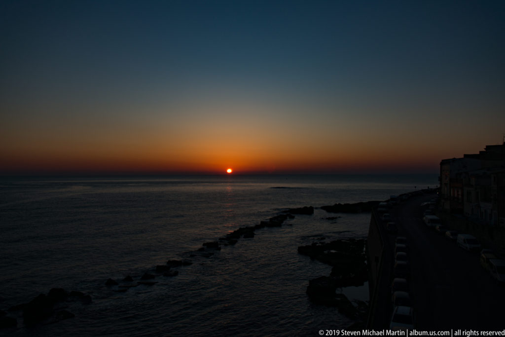 Dark sunset seascape with a small orange sun, silhouetted rocks, and Ortigia’s coastal road at right.
