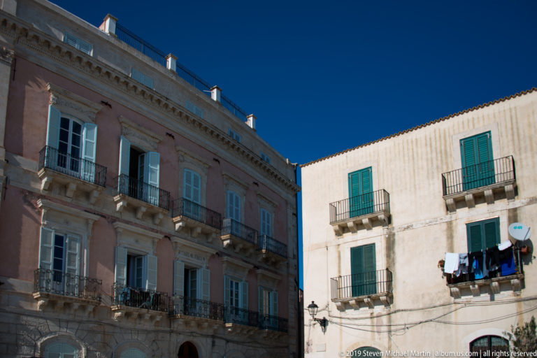 Pink and pale residential buildings with wrought-iron balconies, green shutters, and laundry under a deep blue sky.