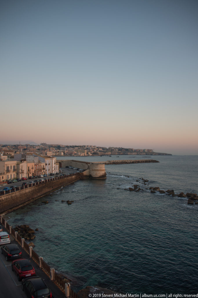 Evening view of a masonry waterfront, road, low buildings, rocky shallows, and distant coastline on Ortigia.