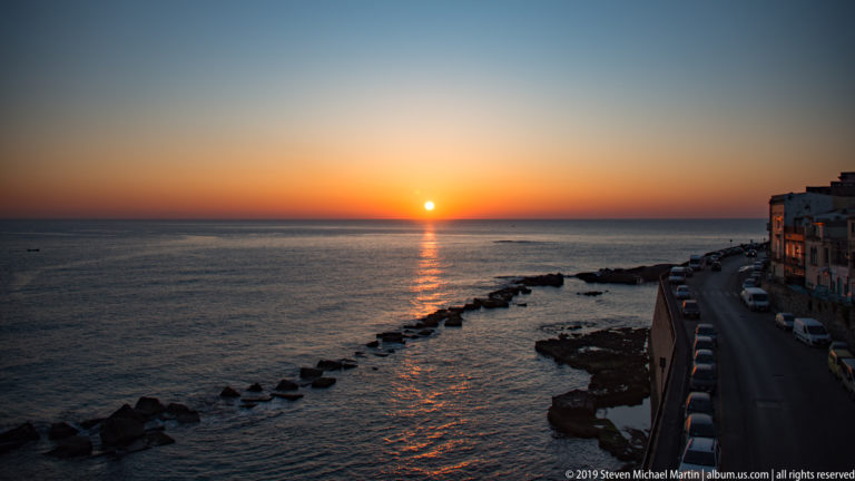 Sunset over the sea beside Ortigia, with a bright reflection, offshore rocks, coastal road, parked cars, and buildings.