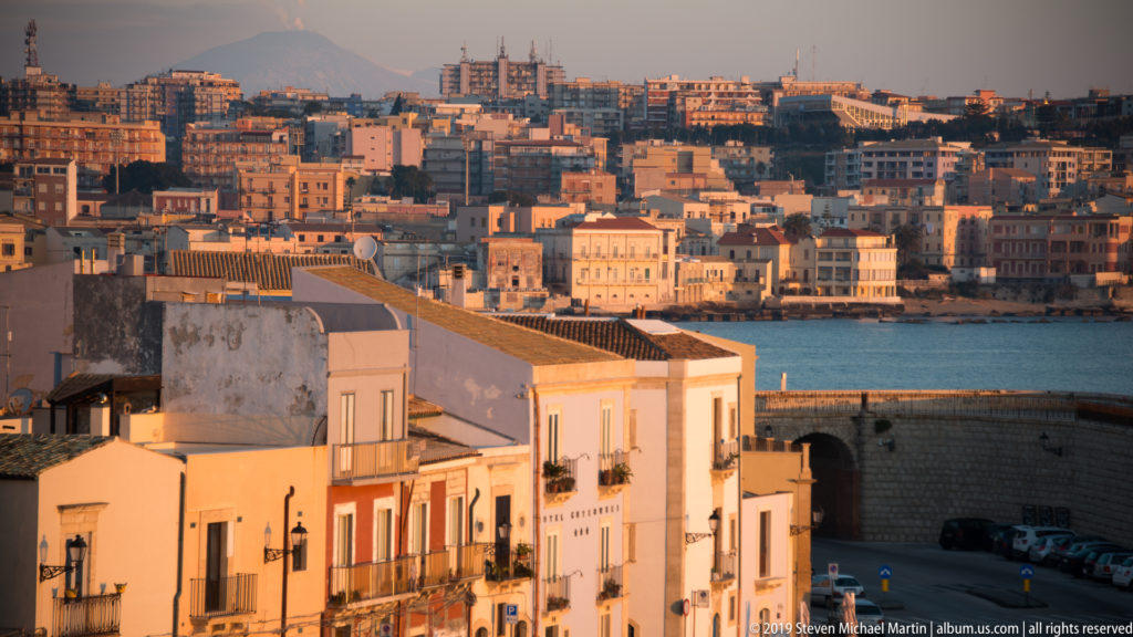Elevated evening cityscape with Ortigia rooftops in front, blue water, and dense Siracusa buildings beyond.