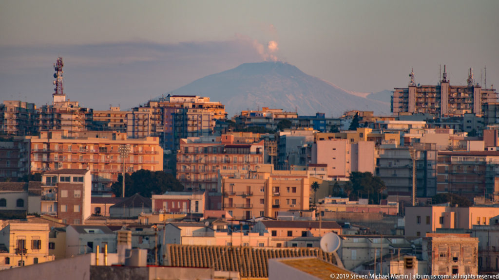 Telephoto cityscape of Siracusa with dense buildings in the foreground and Mount Etna emitting a plume in the distance.