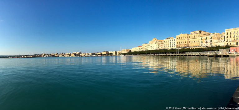 Pale multi-story waterfront buildings on Ortigia reflected in calm blue harbor water beneath a clear sky.