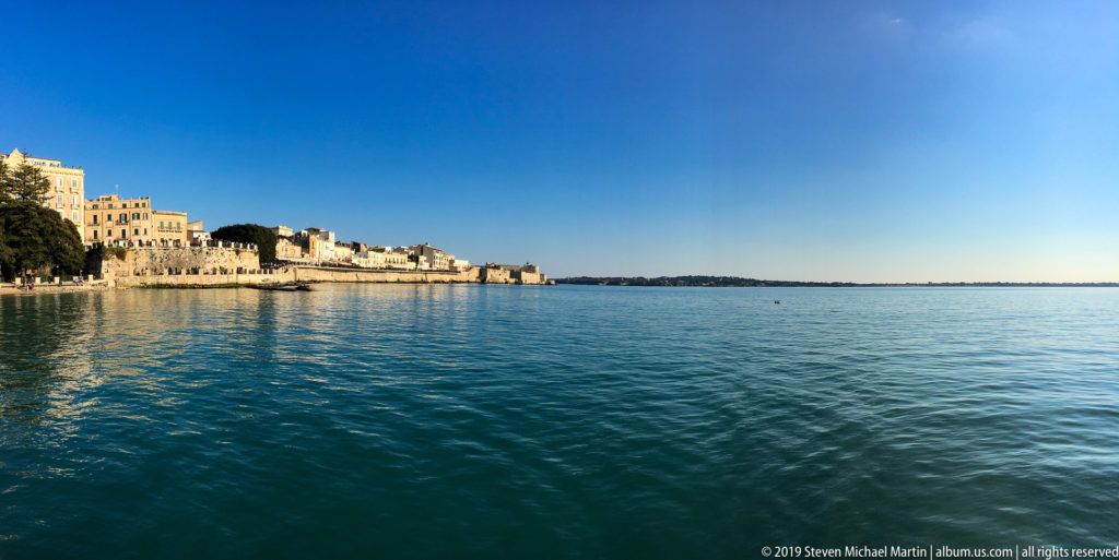 Wide daytime view across calm blue water toward pale historic buildings along Ortigia’s waterfront.