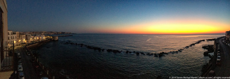 Twilight sea view with orange horizon, offshore rocks, illuminated waterfront buildings, and a seafront road on Ortigia.