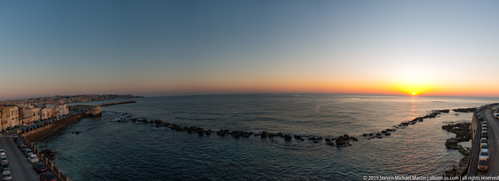 Elevated sunset view over the Ionian Sea, with Ortigia waterfront buildings and a curved line of offshore rocks.