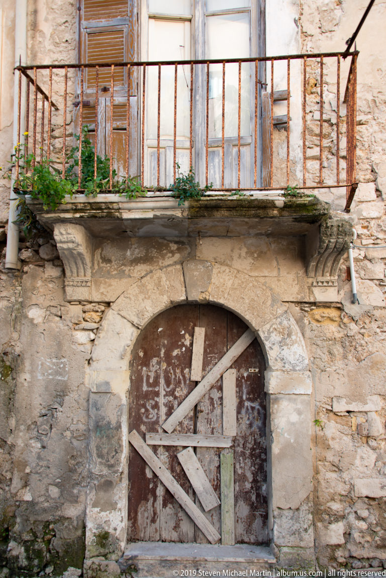 Boarded arched doorway beneath a rusty iron balcony on a heavily weathered stone residential façade.