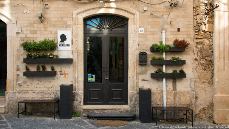 Black Chisessa doorway in an aged stone façade, flanked by rows of wall-mounted planters and low benches.
