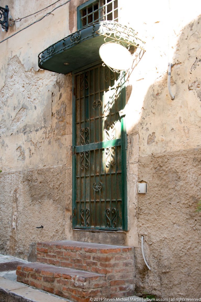 Green metal-grilled doorway above brick steps in a weathered stone wall beneath a small projecting canopy.