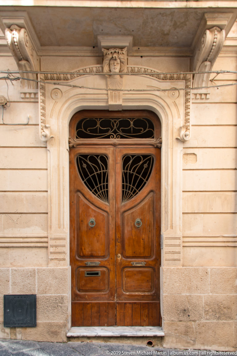 Wooden double door with curving black iron inserts and transom, framed by carved pale stone and a small mask above.