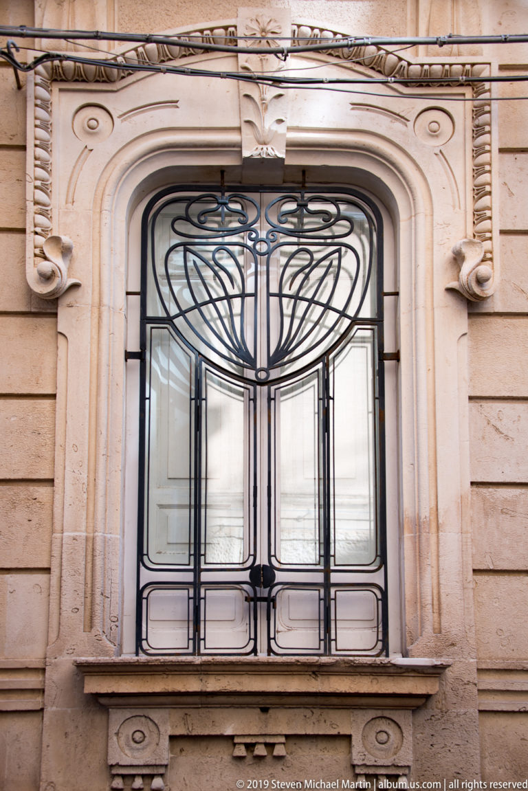 Tall window with symmetrical curving black iron grille framed by ornate pale carved stonework.