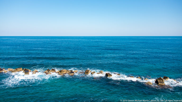 Low line of offshore rocks surrounded by white breaking surf in vivid blue sea beneath a clear horizon.