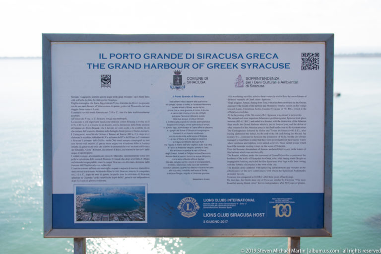 Interpretive sign titled The Grand Harbour of Greek Syracuse standing beside bright blue harbor water.