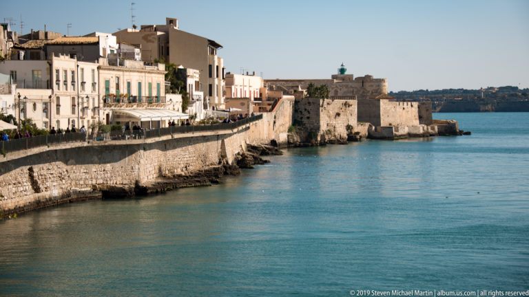 Ortigia waterfront buildings and stone seawall curving toward a fortified projection beside calm blue sea.