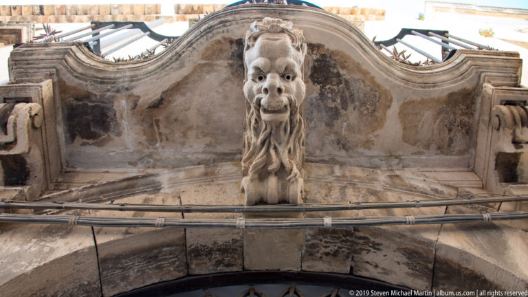 Carved bearded grotesque face projecting from the keystone of a weathered stone arch with cables crossing below.
