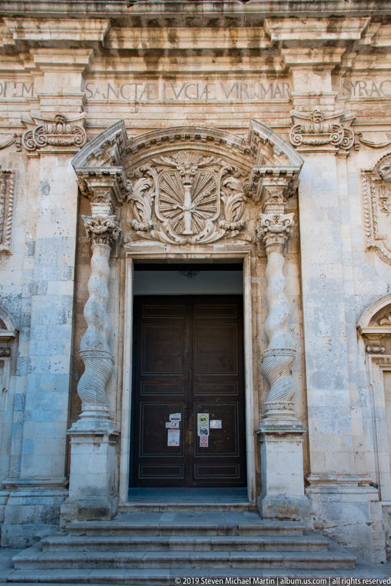 Dark wooden church door reached by steps beneath an ornate stone portal with spiral columns and radiant cross medallion.