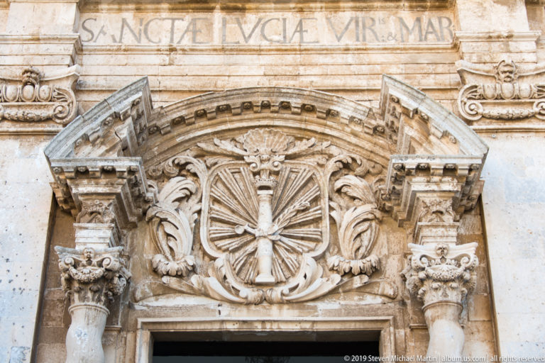 Radiant cross in a circular carved medallion above a doorway, surrounded by leaves, scrolls, columns, and weathered inscription.