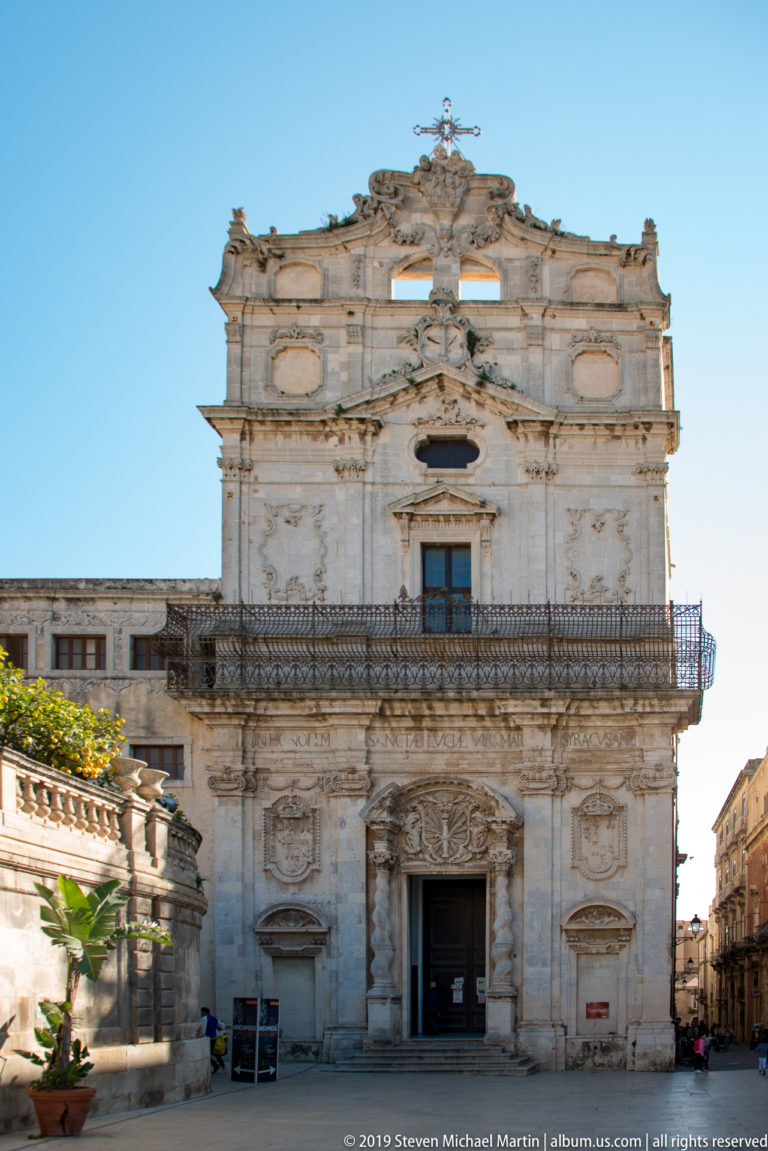Frontal view of an ornate limestone church with carved doorway, iron balcony, oval panels, scrollwork, and crowning cross.