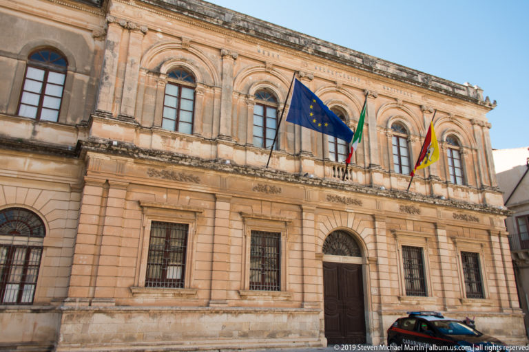 Two-story limestone civic building with arched upper windows and European Union, Italian, and Sicilian flags.