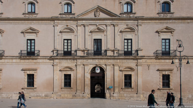 Symmetrical pale-stone palace façade with central arched entrance, pedimented windows, balconies, and a Latin inscription band.