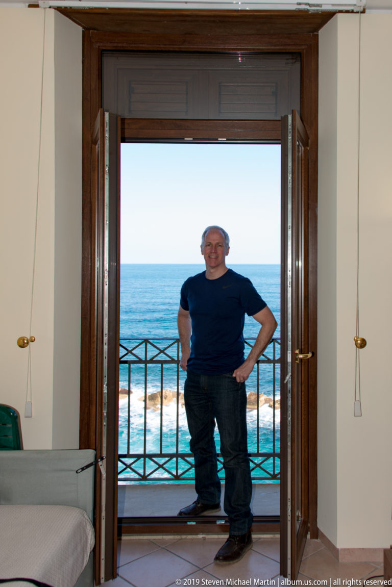 Smiling man standing in open balcony doors with a black railing and bright blue sea directly behind him.