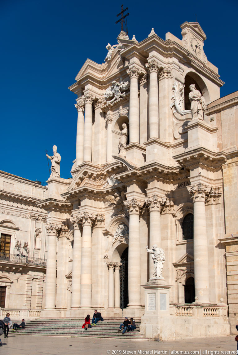 Oblique view of Syracuse Cathedral’s pale Baroque façade with projecting columns, statues, and people in the square.
