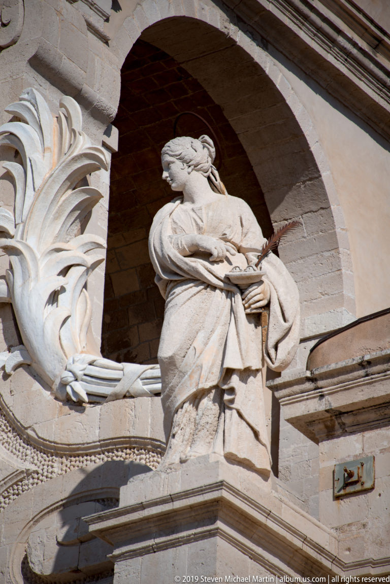 Stone statue of a robed female religious figure in a niche framed by large carved foliage and architectural moldings.