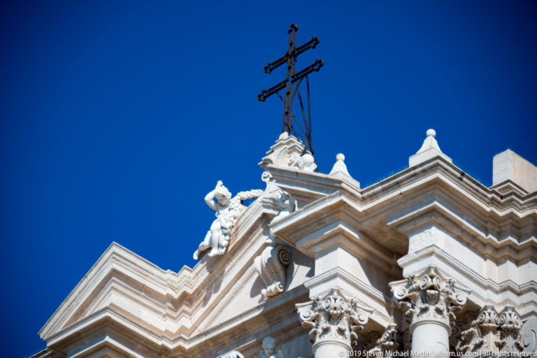 Iron cross above the stepped limestone roofline of Syracuse Cathedral with sculpted figures and Corinthian capitals.