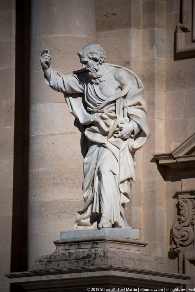 Seated stone male figure in heavy robes extending one arm outward against the pale cathedral wall.