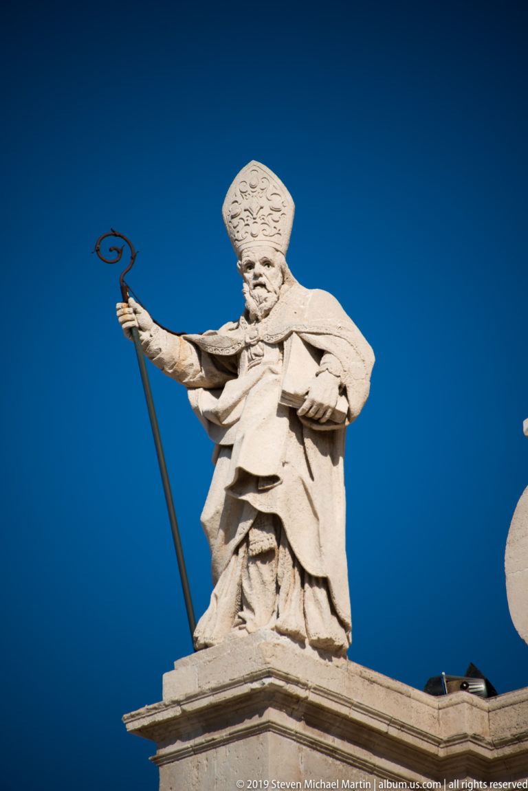 Stone statue of a mitred bishop holding a crozier, silhouetted against a deep blue sky.