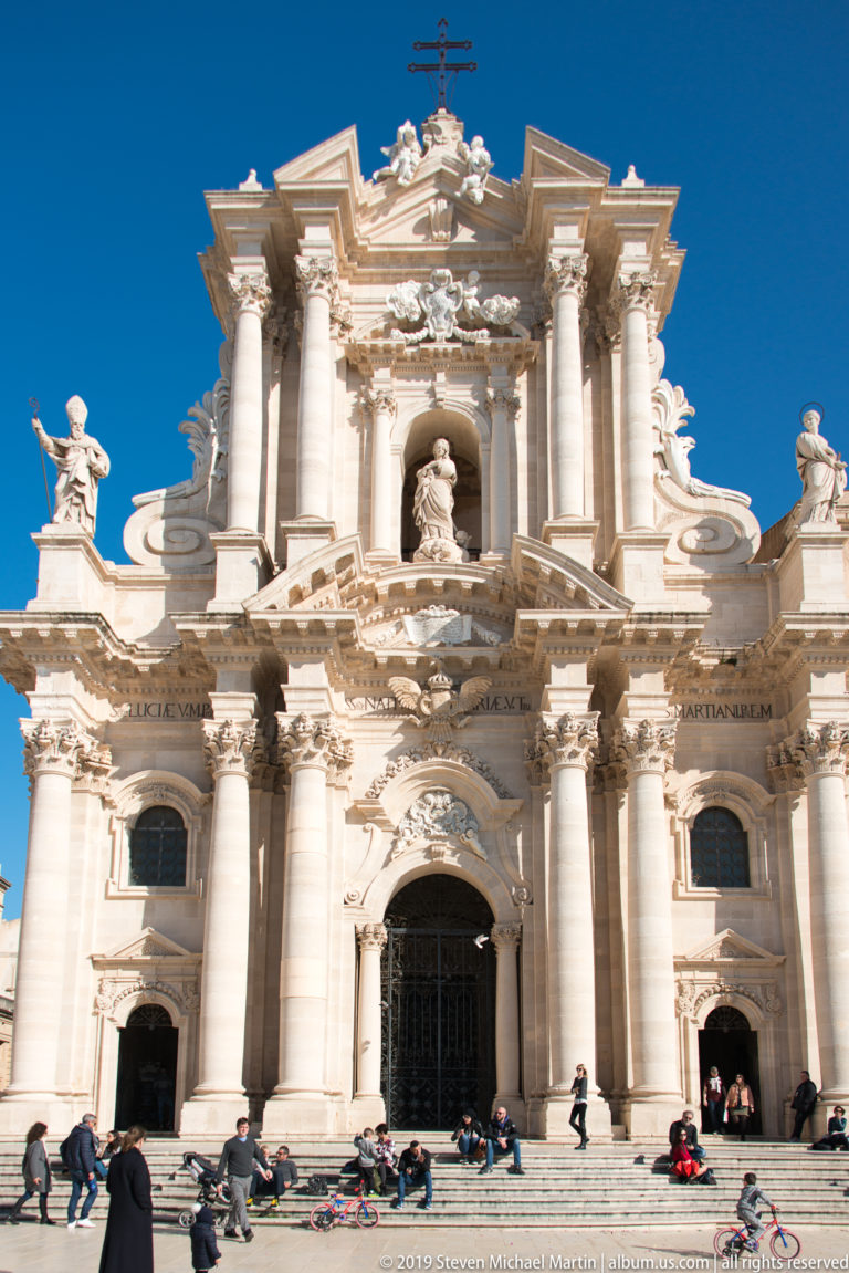 Full frontal view of Syracuse Cathedral’s Baroque façade with columns, statues, broad steps, and people in Piazza Duomo.
