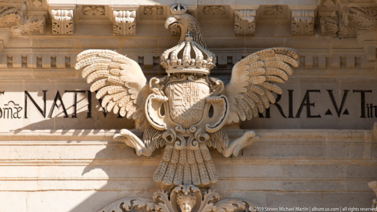 Crowned double-headed stone eagle with shield on its breast, wings spread across an inscribed architectural band.