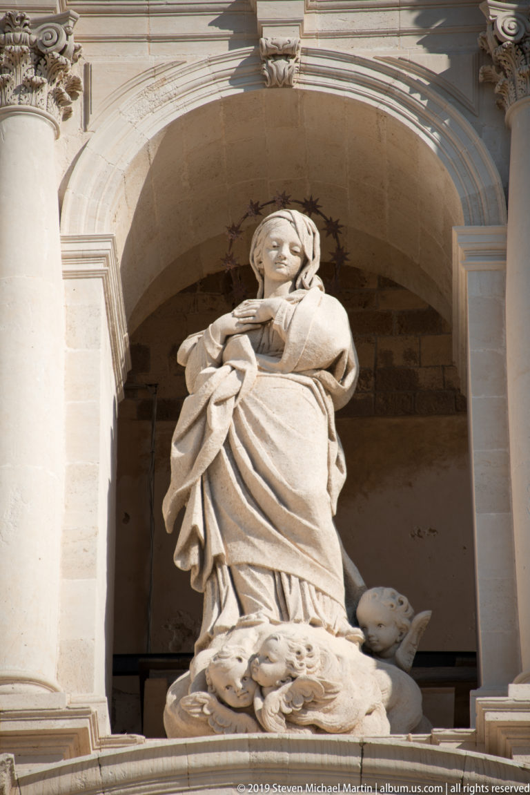 Large robed female saint standing in a deep arched niche on the Syracuse Cathedral façade.