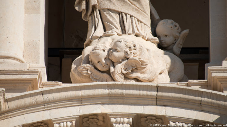 Two stone cherubs with wings nestled against the drapery and base of a larger cathedral statue.