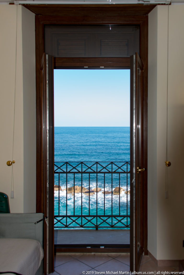 Open wooden balcony doors framing blue sea, black railing, and white surf beyond a simple interior room.