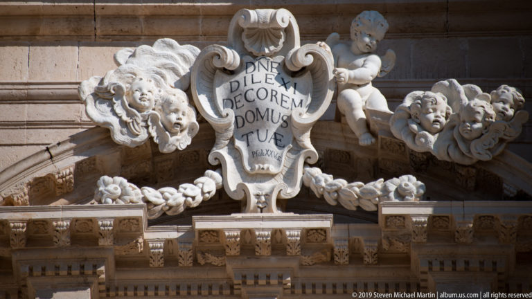 Stone cartouche with Latin inscription surrounded by cherubs, floral swags, and ornate Baroque carving.