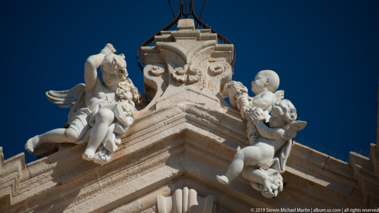 Two carved stone cherubs flanking an ornate heraldic crest high on the cathedral façade against blue sky.