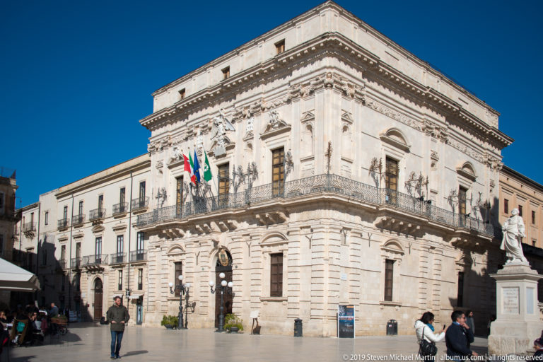 Corner view of Palazzo Vermexio with rusticated ground floor, balcony, civic flags, and adjoining buildings on Piazza Duomo.