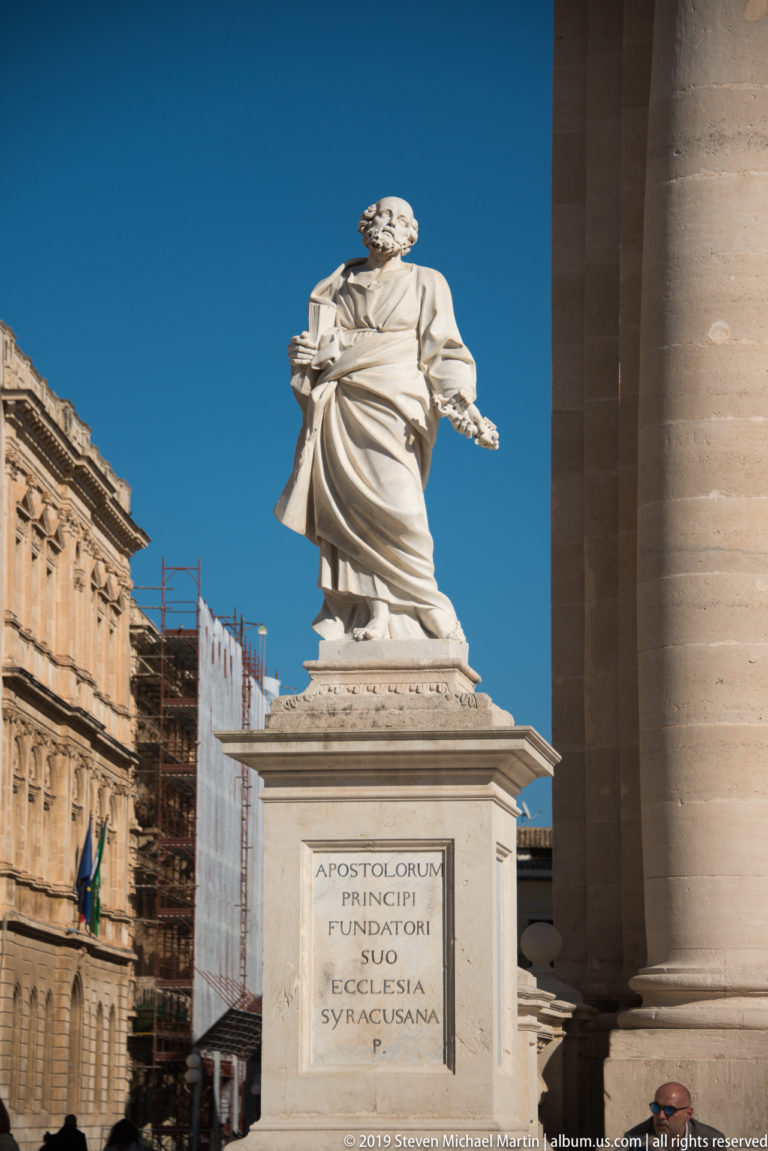 White stone statue of Saint Peter on an inscribed pedestal beside a massive cathedral column in Piazza Duomo.