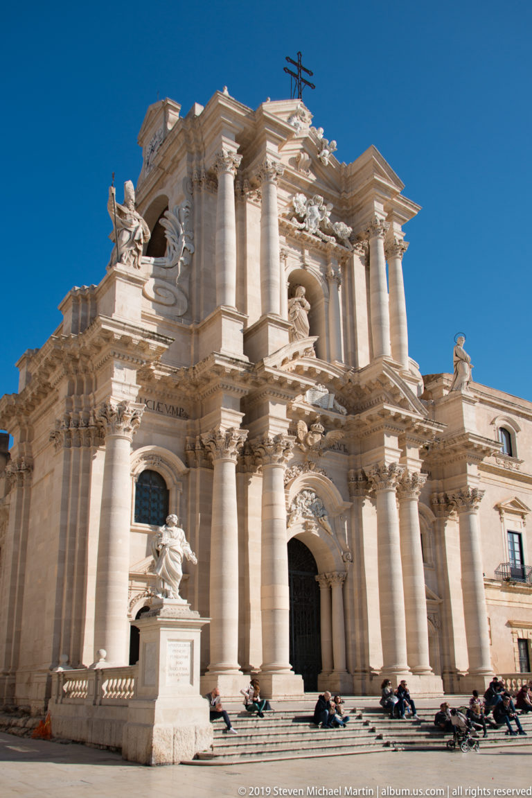 Grand Baroque façade of Syracuse Cathedral with giant columns, statues, and visitors gathered across Piazza Duomo.