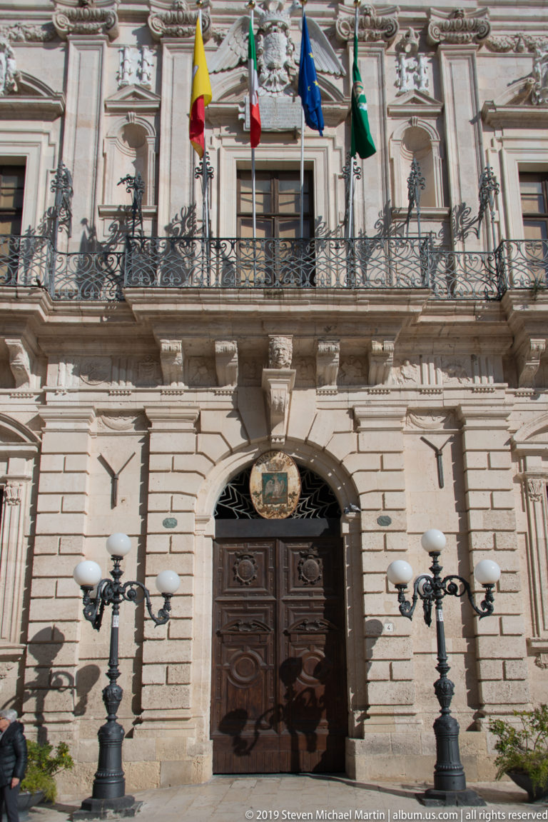 Palazzo Vermexio entrance with rusticated stonework, carved coat of arms, balcony, civic flags, and paired street lamps.