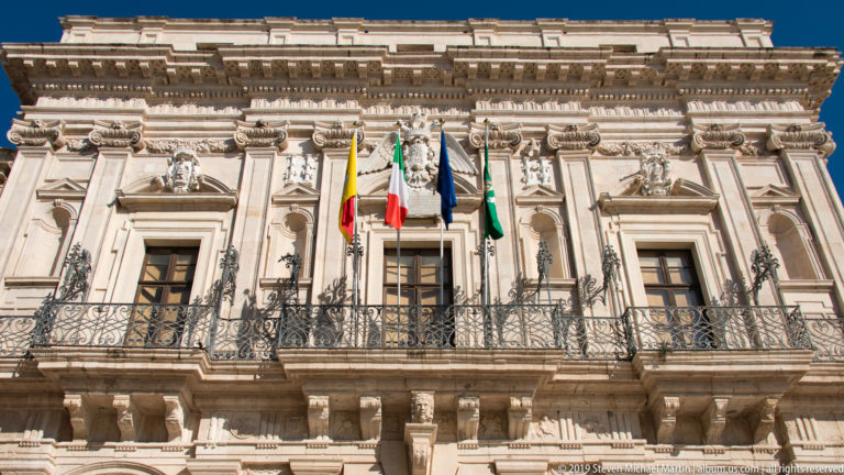 Ornate pale-stone Palazzo Vermexio façade with wrought-iron balcony, carved windows, and four civic flags.