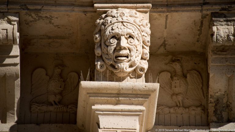 Carved stone grotesque mask with open mouth above shallow relief figures on a historic façade.