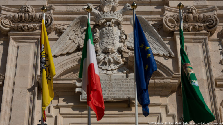 Carved heraldic eagle above a stone tablet, framed by Sicilian, Italian, European Union, and green flags.