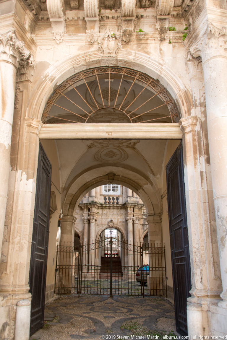 Large arched stone entrance with open wooden doors, fanlight, vaulted passage, and iron gate leading to a courtyard.